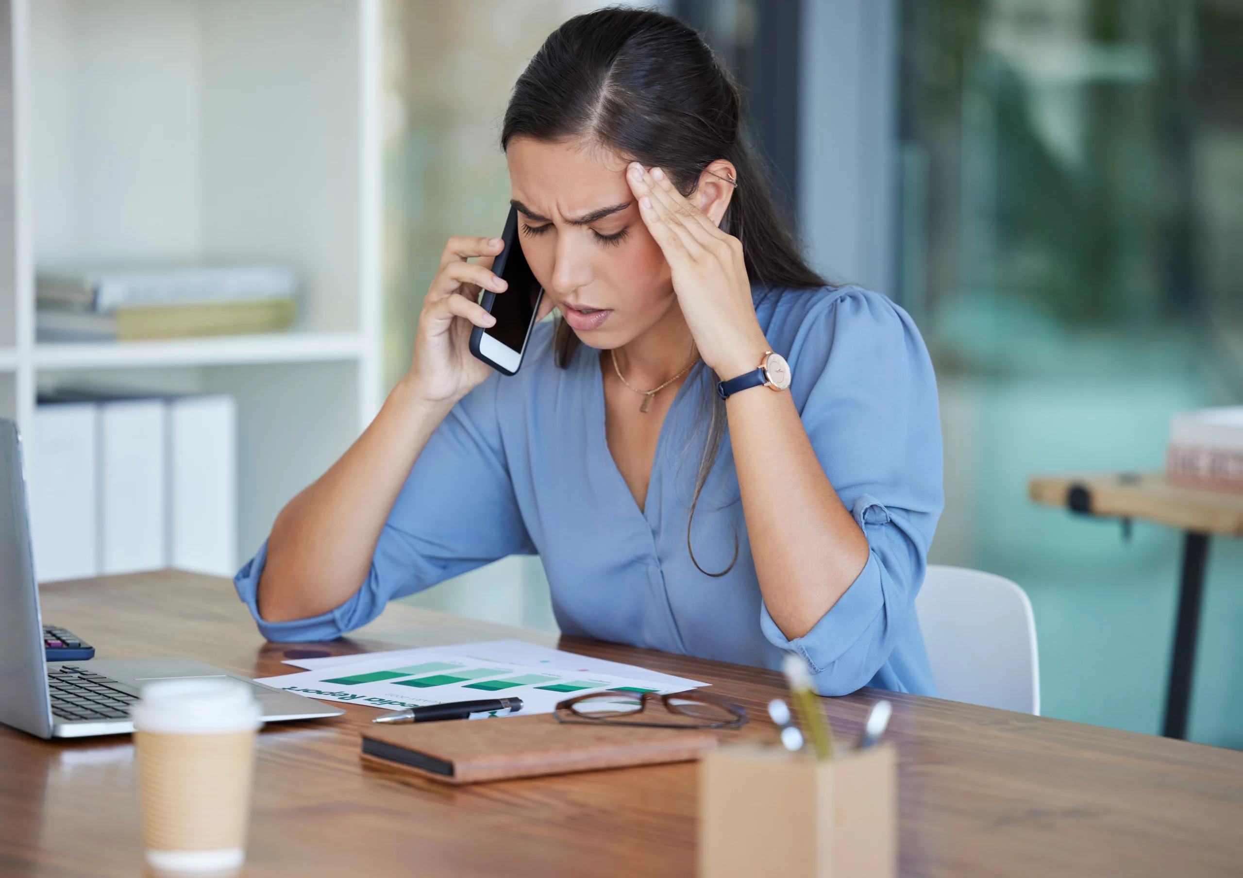 Stressed woman in blue blouse on phone with laptop and documents