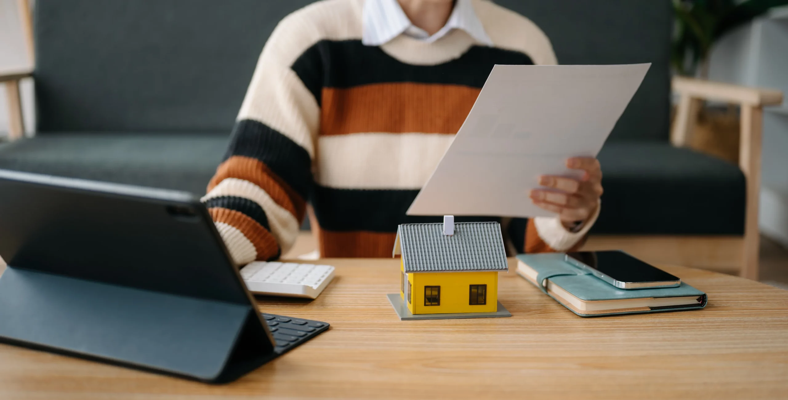 Person in striped sweater at wooden desk with tablet, keyboard, model house, and papers, representing planning to buy a home in Florida after filing bankruptcy and understanding waiting periods